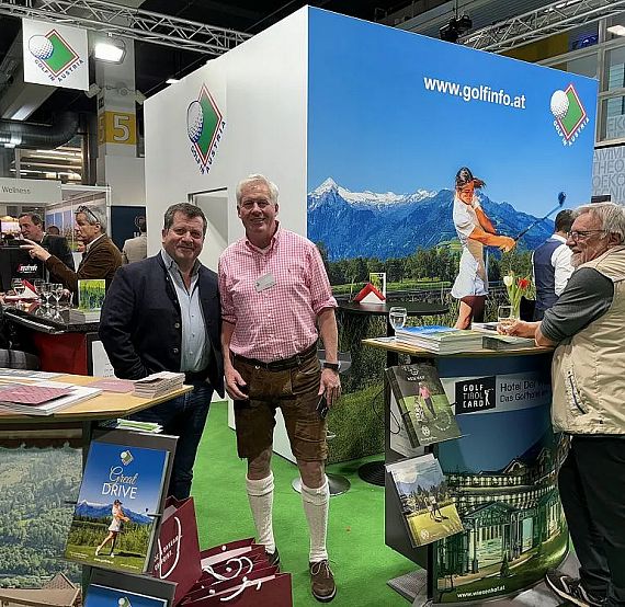 Three men interact at a golf-themed expo booth. A backdrop features mountains and a golfer. Brochures and exhibits are visible, with a casual, friendly atmosphere.