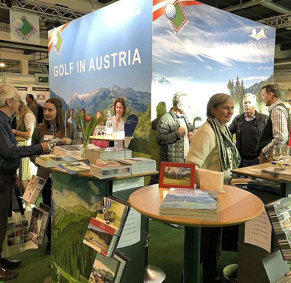 A golf promotion booth with Austrian landscapes, people talking, brochures on the counter, and colorful banners displayed.