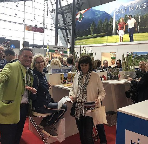 Ein Messe-Stand mit Besuchern, die Informationsmaterial lesen. Eine große Werbetafel im Hintergrund zeigt „Golf in Austria“. Menschen unterhalten sich angeregt.