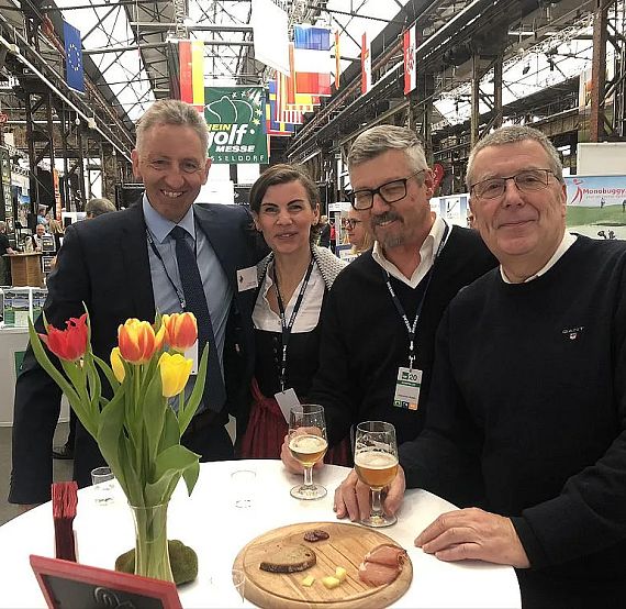 Four people at a table in an industrial venue, enjoying drinks and snacks, with tulips in a vase, under overhead lights and flags.