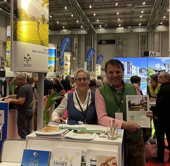 People at a trade fair booth with promotional displays, surrounded by convention attendees.