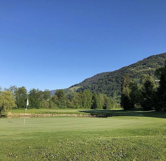 View of a golf course green with a flag, surrounded by trees and a hill under a clear blue sky. The scene is peaceful and well-maintained.