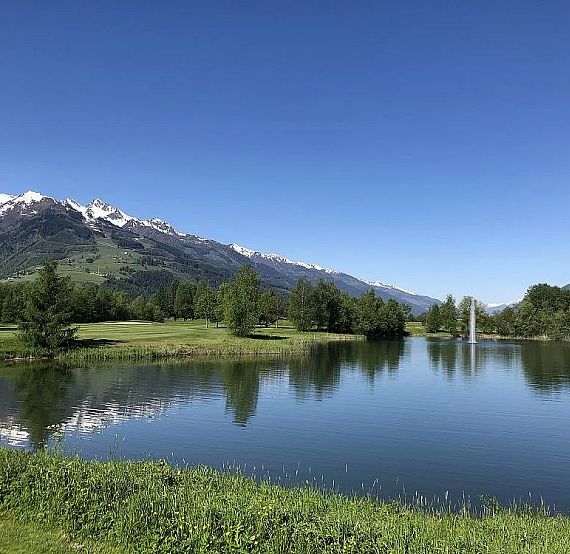 A tranquil lake surrounded by green fields and trees, with snow-capped mountains in the background under a clear blue sky.