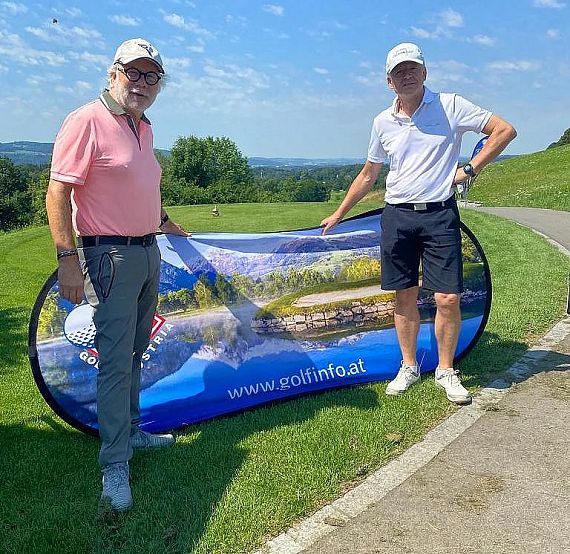 Zwei Männer stehen auf einem Golfplatz neben einem Banner. Einer trägt ein rosa Poloshirt, der andere ein weißes Poloshirt. Hügelige Landschaft im Hintergrund.
