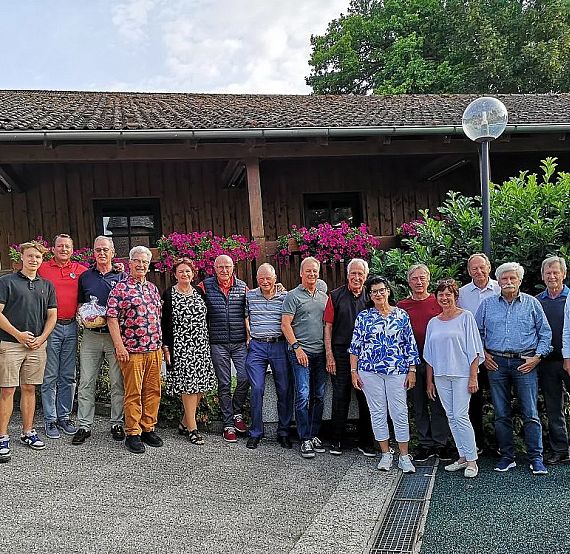 A group of adults posing outside in front of a building with wooden walls and pink flowers. They are casually dressed and smiling in the daylight.