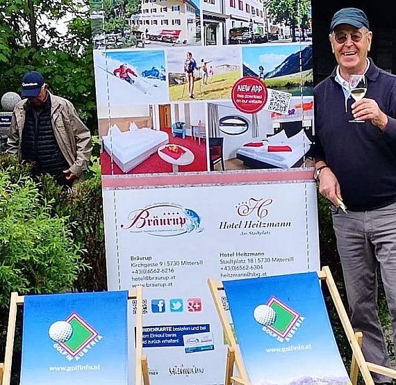 Man in a hat smiles, holding a drink, near hotel marketing signage and deck chairs at an outdoor setting with greenery.