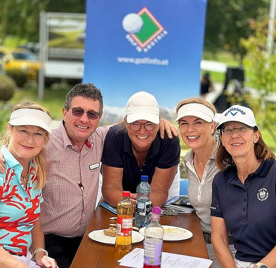 Fünf Personen, vier Frauen und ein Mann, lächeln bei einem Tisch im Freien. Sie tragen Golfkleidung, und im Hintergrund ist ein Golfplatz-Banner zu sehen.