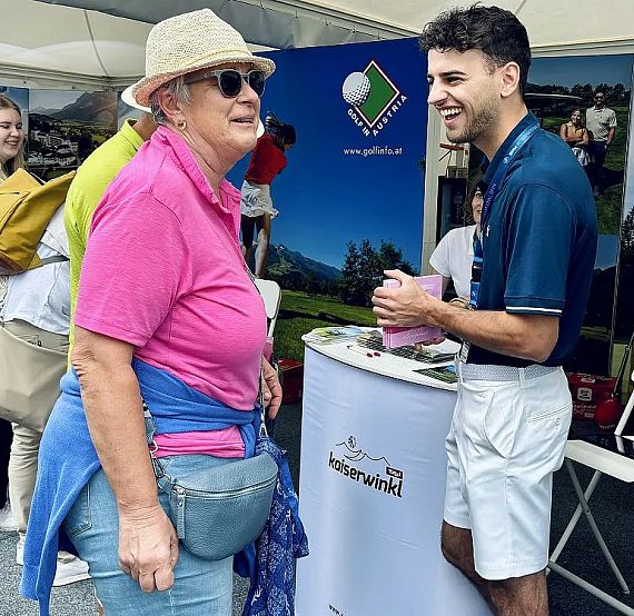 Two people smiling at a booth, possibly at an outdoor event. One person wears a hat and pink shirt, the other in a polo shirt and shorts.
