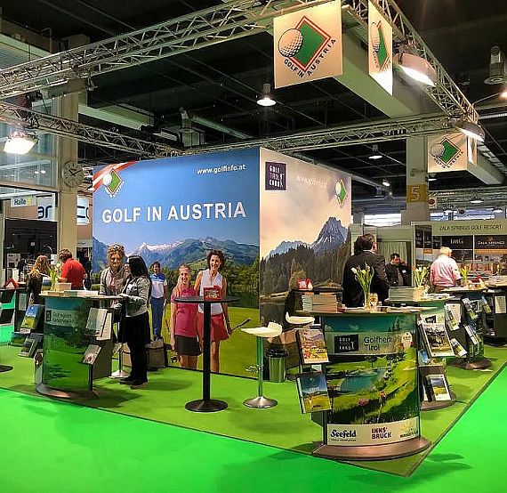 Trade fair booth showcasing Austrian golf tourism with banners, brochures, and people interacting, set on a green carpet to mimic a golf environment.