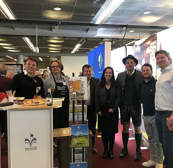 A group of people standing indoors at an exhibition booth with brochures and products on display. They are smiling and dressed in casual to formal attire.