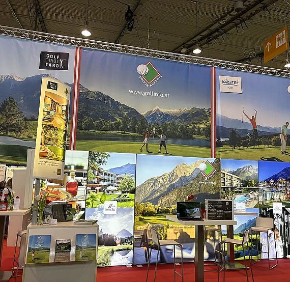 Trade show booth with colorful banners depicting mountains and outdoor activities, promoting tourism. Tables and display stands are arranged on a red carpet.