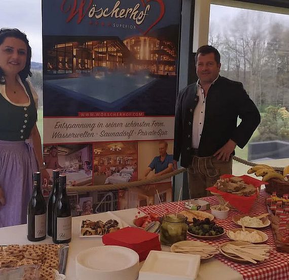 A man and woman in traditional Bavarian attire stand by a food table with sausages, pretzels, wine, and promotional banner in a rustic setting.