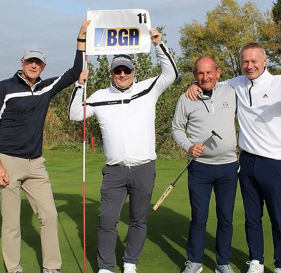 Four men on a golf course wearing casual golf attire, holding a flag with 'BGA' on it. They are smiling and standing on the green under a clear sky.