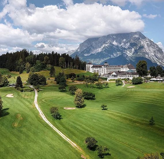 Lush green landscape with a winding path, scattered trees, and a large building, set against majestic mountains under a partly cloudy sky.