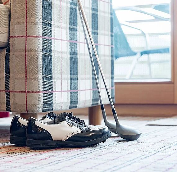 Black and white golf shoes on a rug by a checkered armchair, with golf clubs leaning against it, and outdoor furniture visible through a window.