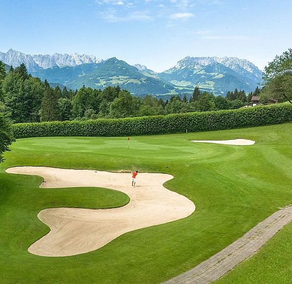 A golf course with green fairways, sand bunkers, and alpine mountains in the background under a clear blue sky.