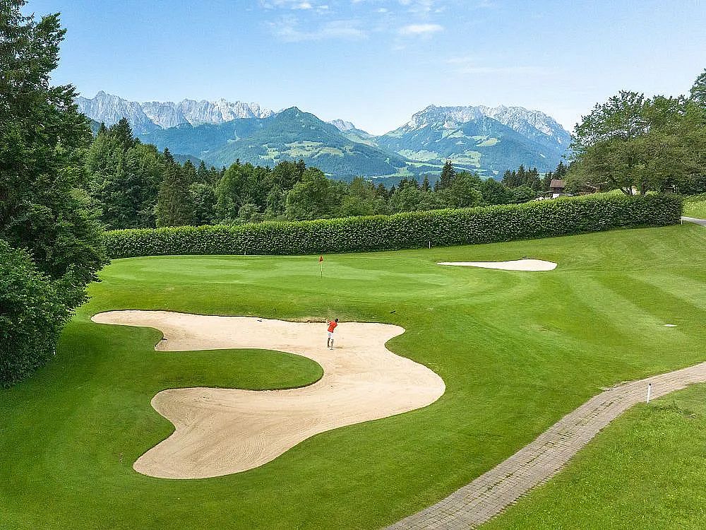 A golf course with green fairways, sand bunkers, and alpine mountains in the background under a clear blue sky.