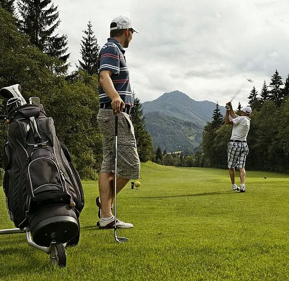Two men are on a golf course. One swings a club, while the other stands nearby with a golf bag. The course is surrounded by trees and mountains.
