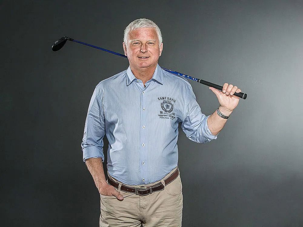 A man in a light blue shirt with rolled-up sleeves, posing with a golf club on his shoulder. He stands in front of a neutral gray background, smiling.