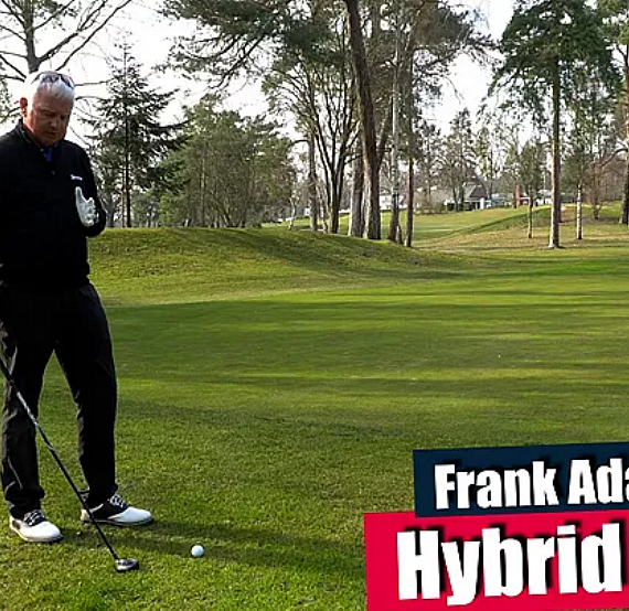 A man in a golf course setting is preparing to swing a club. He stands on a green lawn with trees in the background and a golf cart nearby.