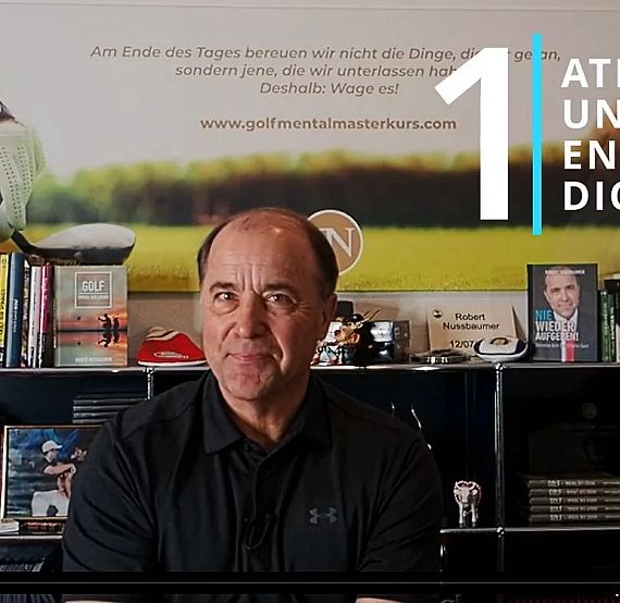 A man in a black shirt sits in front of a bookshelf filled with books and golf memorabilia. A motivational golf poster is displayed behind him.