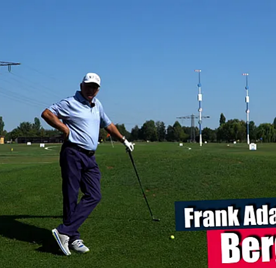 Golfer in blue shirt and white cap holding club on sunny driving range, with tall structures and trees in background, clear blue sky above.