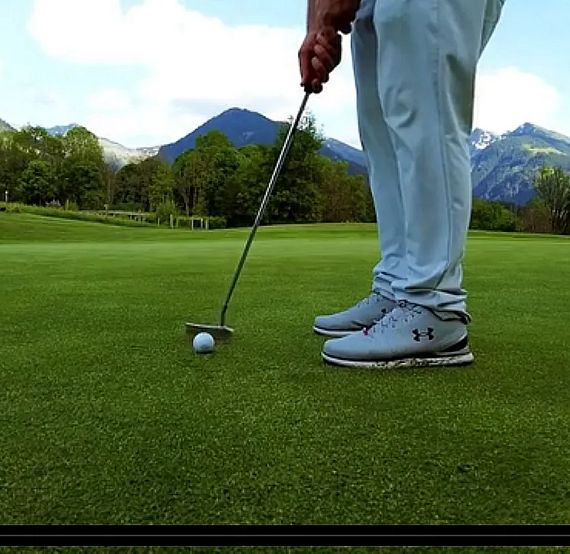 A golfer putting on a green with mountains in the background, wearing light-colored sportswear and shoes.