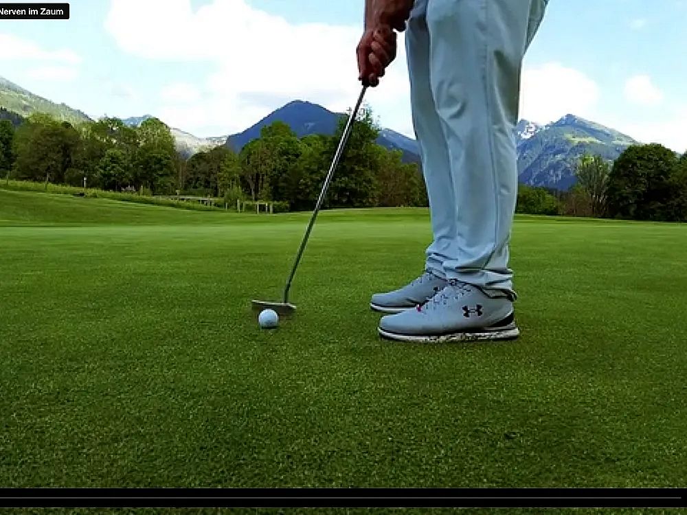 A golfer putting on a green with mountains in the background, wearing light-colored sportswear and shoes.