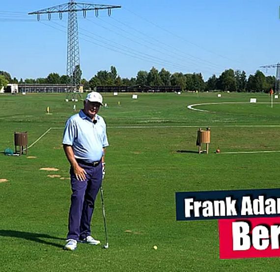 A golfer on a driving range under a blue sky, surrounded by practice golf balls and trees in the background.