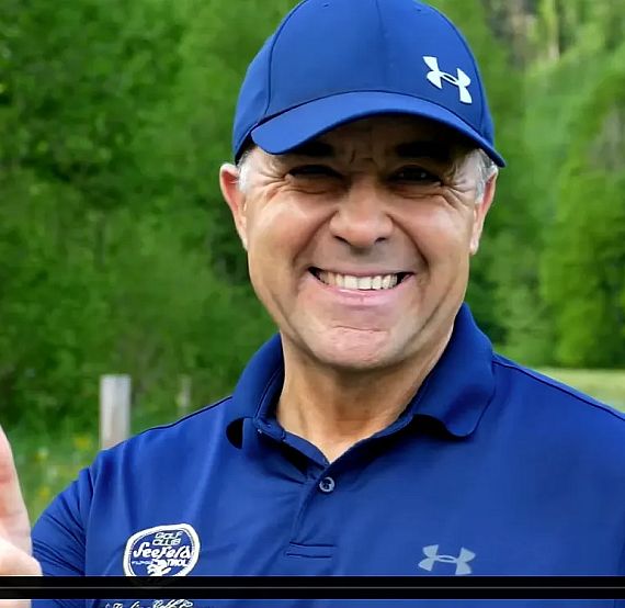 A smiling person outdoors wearing a blue cap and polo shirt, giving a thumbs-up. Greenery is in the blurred background, evoking a casual atmosphere.