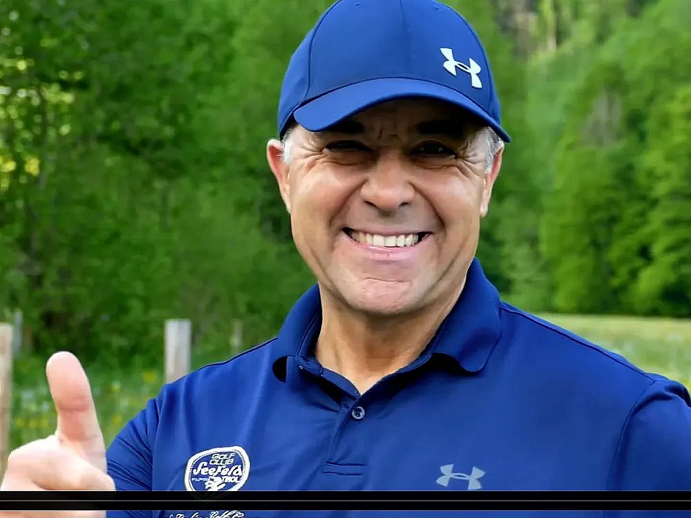 A smiling person outdoors wearing a blue cap and polo shirt, giving a thumbs-up. Greenery is in the blurred background, evoking a casual atmosphere.