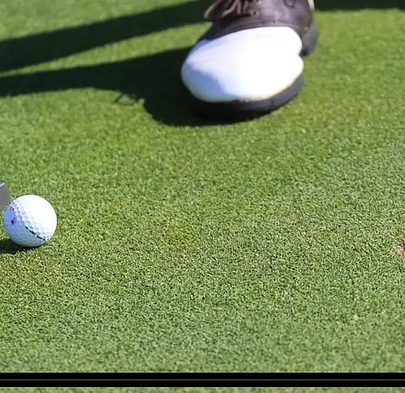 Close-up of a golfer's putter and ball on a green, with the ball near a hole. A shoe is visible, indicating a motion to strike the ball.