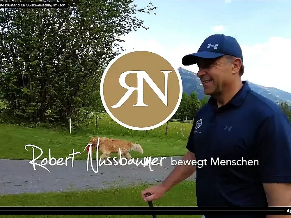Ein Mann mit Baseballmütze und Golfschläger geht lächelnd über einen Weg in einer grünen Parklandschaft. Im Hintergrund sind Bäume und Berge sichtbar.