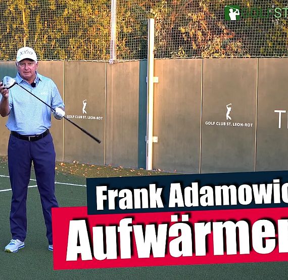 A man standing on a tennis court in front of a fence holding a golf club, with trees in the background.