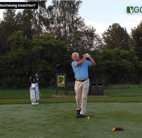 A golfer in mid-swing on a lush golf course, surrounded by trees and using a large bag of clubs, practicing his technique.