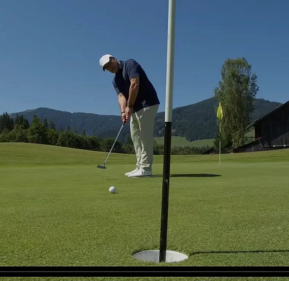 Golfer putting on a green under clear blue sky, with mountains and a barn in the background. The scene is peaceful and sunny, ideal for a day of golf.