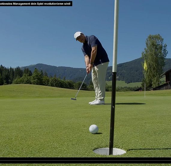 A golfer in a blue shirt and white cap putts the ball on a sunny day with hills in the background.
