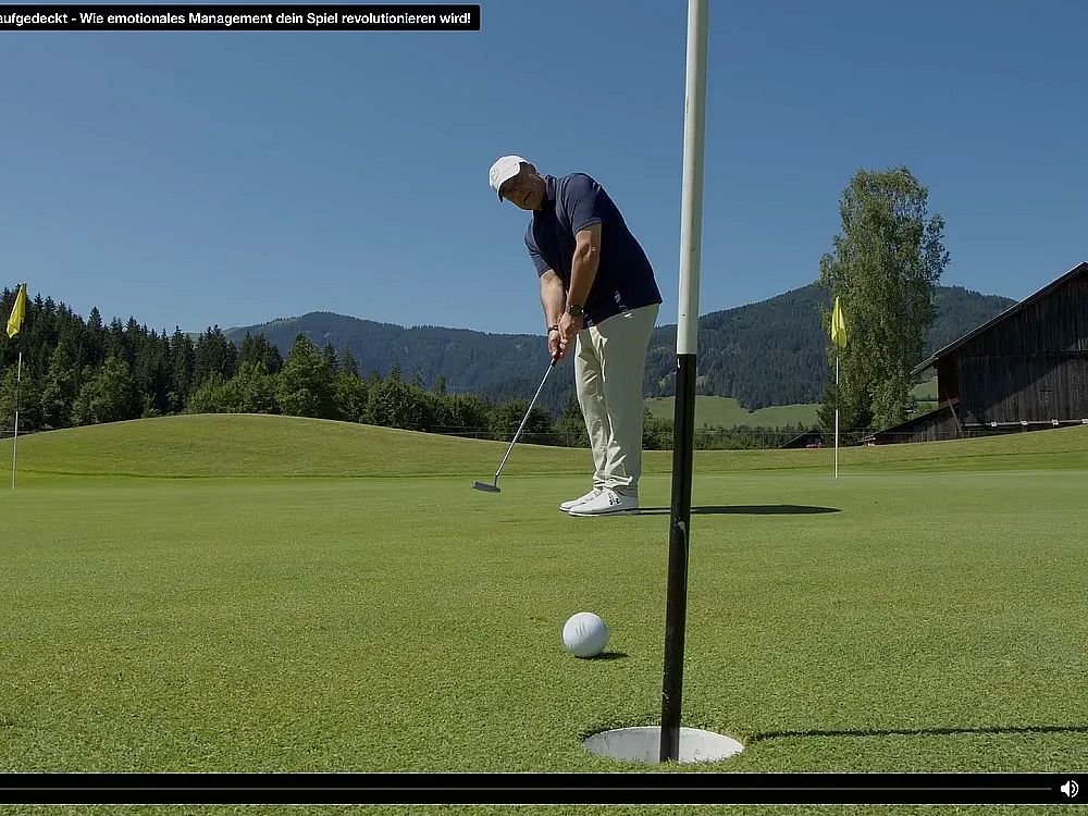 A golfer in a blue shirt and white cap putts the ball on a sunny day with hills in the background.