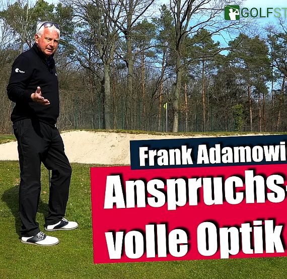 A man on a golf course next to a bunker, gesturing with hand, trees in background, clear weather.