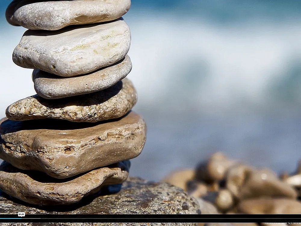 A stack of smooth, flat stones is balanced on a rocky beach, with a blurry ocean in the background, creating a peaceful and serene atmosphere.