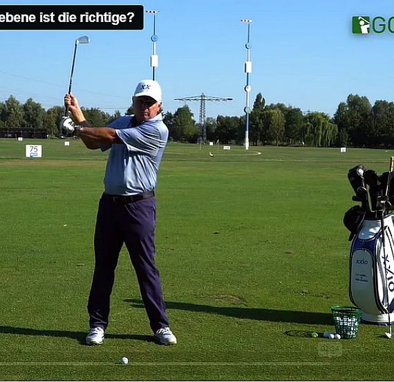 A golfer demonstrating a swing on a green course, with a golf bag and clubs nearby under a clear sky.