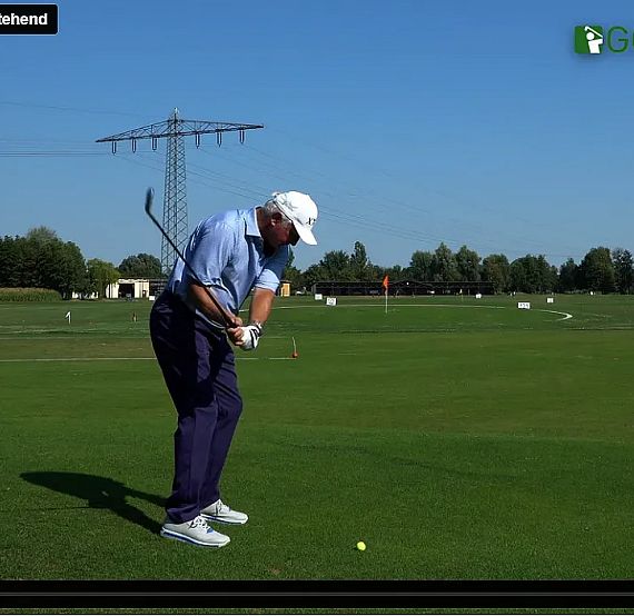 Man golfing on a green field under a blue sky, positioned mid-swing with trees and a power line visible in the distance.
