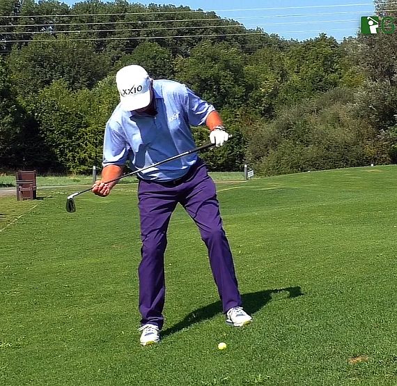 A golfer in a light blue shirt and dark pants prepares for an uphill shot on a green golf course surrounded by trees.