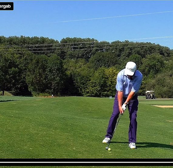 Golfer in mid-swing on a green golf course with trees in the background, wearing a white cap and light blue shirt.
