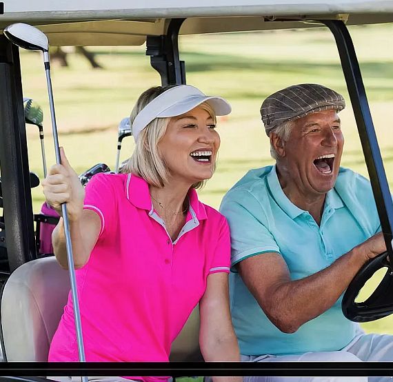 Elderly couple in a golf cart, woman holding a club, both laughing in sporty attire. Sunny golf course background.