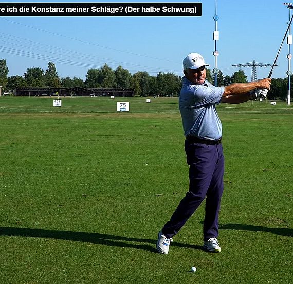 A golfer practices a half-swing on a green golf course with trees in the background under a clear blue sky.