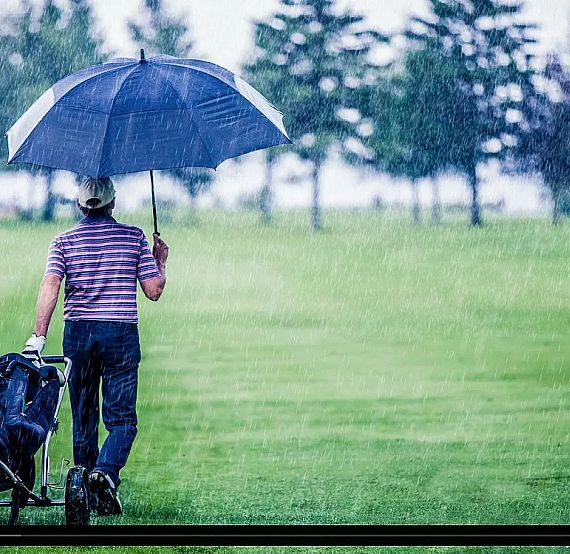 Golfer with umbrella on a rainy course, pulling a golf bag, with trees in the distance, creating a serene yet wet scene.