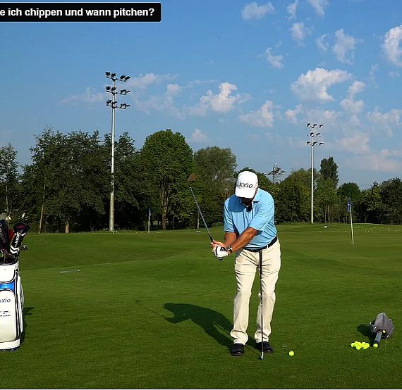 A golfer in blue shirt and white pants prepares to hit a golf ball on a green field under a clear blue sky.