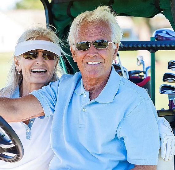 An elderly couple sits in a golf cart on a sunny day, smiling and wearing sunglasses, with golf clubs in the back. They appear relaxed and happy.
