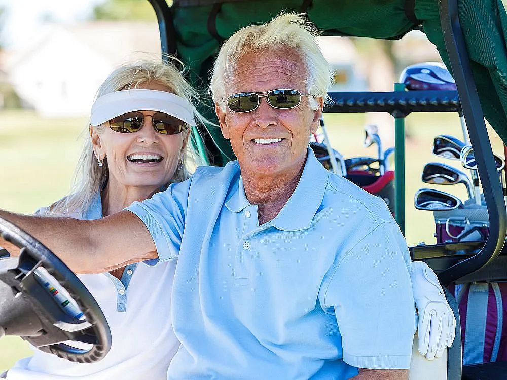 An elderly couple sits in a golf cart on a sunny day, smiling and wearing sunglasses, with golf clubs in the back. They appear relaxed and happy.
